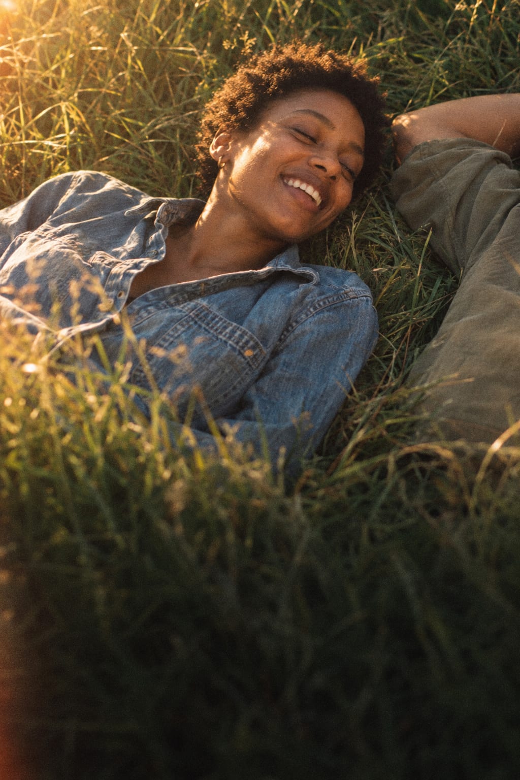 A young woman with short natural hair lying in long grass at golden hour beside her partner, eyes closed, smiling.