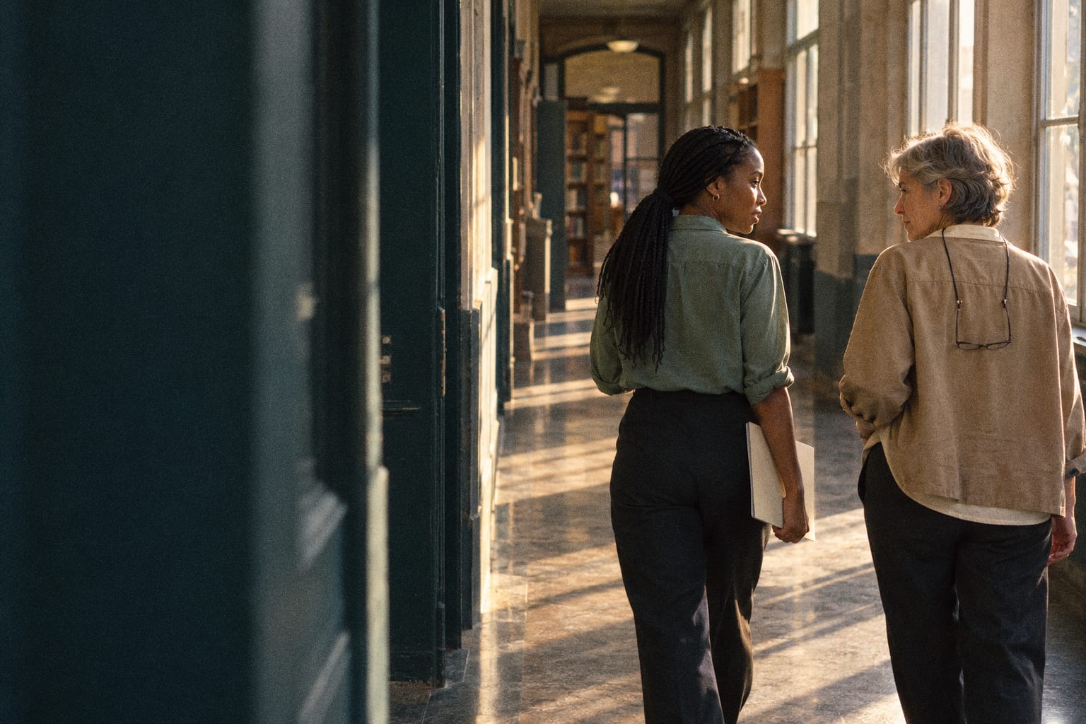 Two colleagues walking and talking down a sunlit corridor, a younger woman with long braids and an older woman with short grey hair.