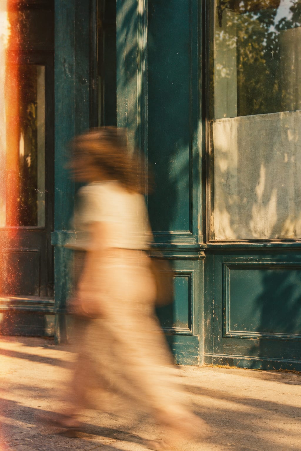 A woman walking past a deep teal shopfront in late sun, blurred by motion, a warm light leak at the edge of the frame.